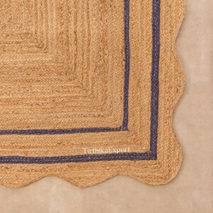 Puede incluir: Una alfombra de yute natural con borde festoneado y borde azul oscuro. La alfombra tiene una textura tejida y una forma rectangular con un borde festoneado. La alfombra es de color neutro y sería adecuada para una sala de estar o un dormitorio.