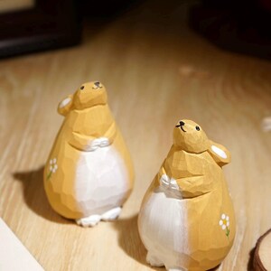 May include: Two wooden rabbit figurines. The rabbits are carved with a yellow-brown body and white belly. Each has a small white flower painted on the side. The rabbits are posed with their paws clasped.