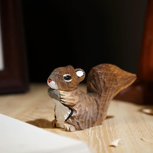 May include: A hand-carved wooden squirrel figurine with a detailed, textured finish. The squirrel is brown with a white belly, black and white eyes, and a red nose. It appears to be holding a nut. The figurine is on a wooden surface.