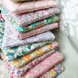 May include: A stack of rectangular quilted pouches featuring various floral patterns in shades of pink, yellow, blue, green, and lavender. Each pouch has a diamond-stitched texture and a soft fabric finish, arranged in a vertical pile.