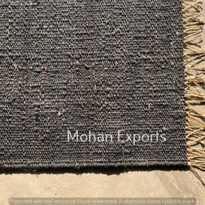 May include: Close-up of a dark gray woven rug with a textured surface and a natural fringe edge. The rug has the text "Mohan Exports" in white. The rug is made of a durable material.