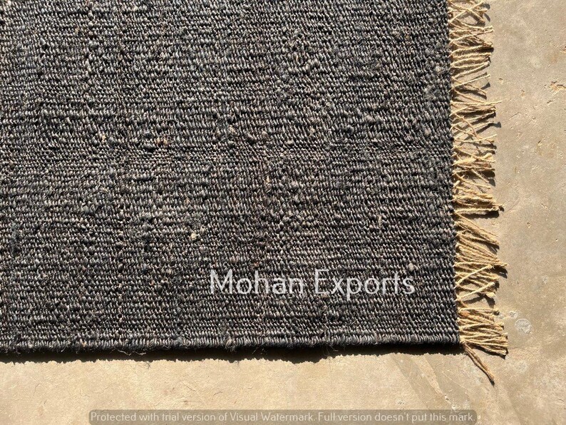 May include: Close-up of a dark gray woven rug with a textured surface and a natural fringe edge. The rug has the text "Mohan Exports" in white. The rug is made of a durable material.