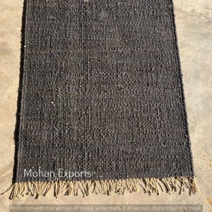 May include: A rectangular, dark gray jute rug with a textured weave. The rug has a natural, fringed edge and the text "Mohan Exports" is visible. The rug is displayed on a concrete surface.