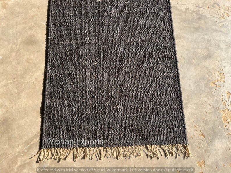 May include: A rectangular, dark gray jute rug with a textured weave. The rug has a natural, fringed edge and the text "Mohan Exports" is visible. The rug is displayed on a concrete surface.