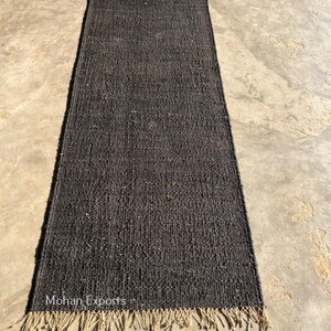 May include: A rectangular, dark gray jute rug with a fringed edge. The rug has a woven texture and is displayed on a concrete surface. The text "Mohan Exports" is visible on the rug.