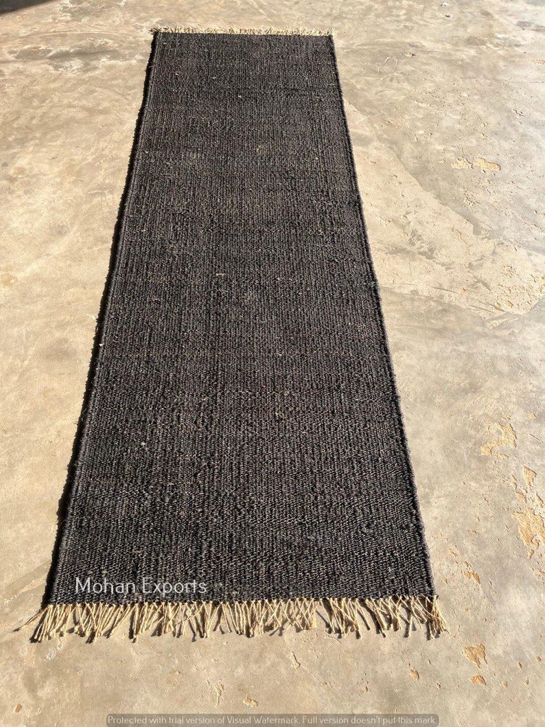 May include: A rectangular, dark gray jute rug with a fringed edge. The rug has a woven texture and is displayed on a concrete surface. The text "Mohan Exports" is visible on the rug.