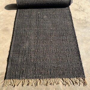 May include: A rolled-up, rectangular, dark gray rug with a woven texture. The rug has a fringe of natural-colored fibers along one edge. The rug is unrolled on a concrete surface, with the rolled portion at the top of the image.