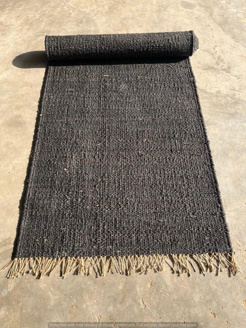 May include: A rolled-up, rectangular, dark gray rug with a woven texture. The rug has a fringe of natural-colored fibers along one edge. The rug is unrolled on a concrete surface, with the rolled portion at the top of the image.
