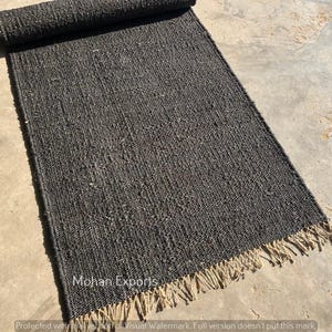May include: A dark gray rectangular rug with a rolled-up section at the top. The rug has a woven texture and a fringe along the bottom edge. The rug is on a concrete surface. The text "Mohan Exports" is visible.