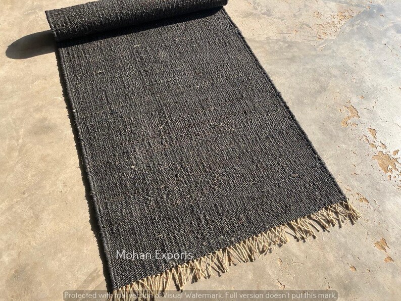 May include: A dark gray rectangular rug with a rolled-up section at the top. The rug has a woven texture and a fringe along the bottom edge. The rug is on a concrete surface. The text "Mohan Exports" is visible.