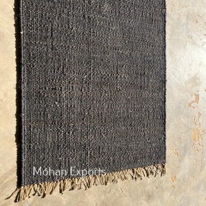 May include: A rectangular, dark gray woven rug with a fringed edge. The rug has a textured appearance and is made of a natural fiber. The text "Mohan Exports" is visible on the rug. The rug is displayed on a concrete surface.