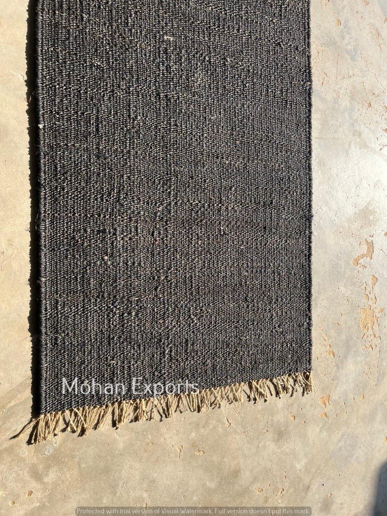 May include: A rectangular, dark gray woven rug with a fringed edge. The rug has a textured appearance and is made of a natural fiber. The text "Mohan Exports" is visible on the rug. The rug is displayed on a concrete surface.