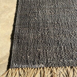 May include: A close-up of a dark gray woven rug with a textured surface and a natural fiber fringe. The rug has the text "Mohan Exports" on it.