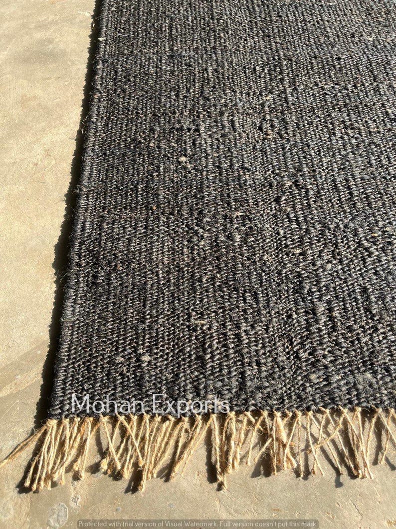 May include: A close-up of a dark gray woven rug with a textured surface and a natural fiber fringe. The rug has the text "Mohan Exports" on it.