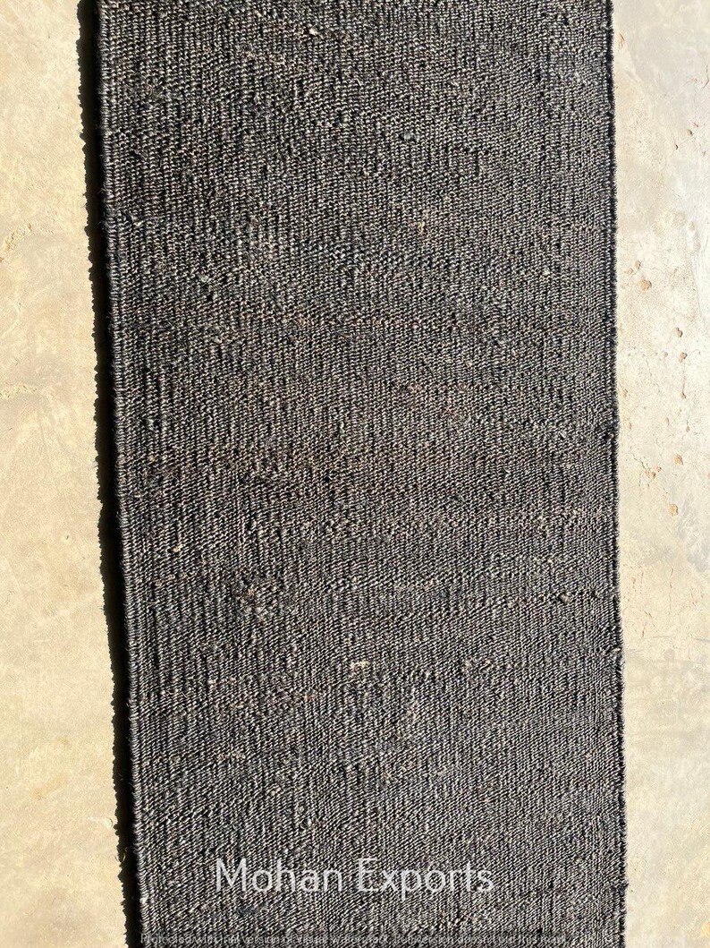 May include: A rectangular, dark gray woven rug. The rug has a textured surface and is made of a natural fiber. The words "Mohan Exports" are printed in white at the bottom of the rug. This rug is suitable for home decor.