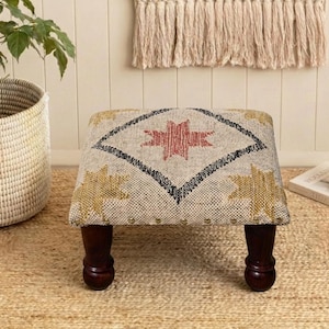 May include: A small, upholstered footstool with a geometric design in red, black, and gold. The stool has dark brown, rounded legs and is placed on a woven jute rug. A woven basket with a plant is to the left.