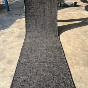 May include: A long, rectangular charcoal gray rug with a woven texture. The rug has a natural fiber fringe at the bottom edge. The rug is displayed outdoors on a concrete surface. The text "Mohan Exports" is visible.