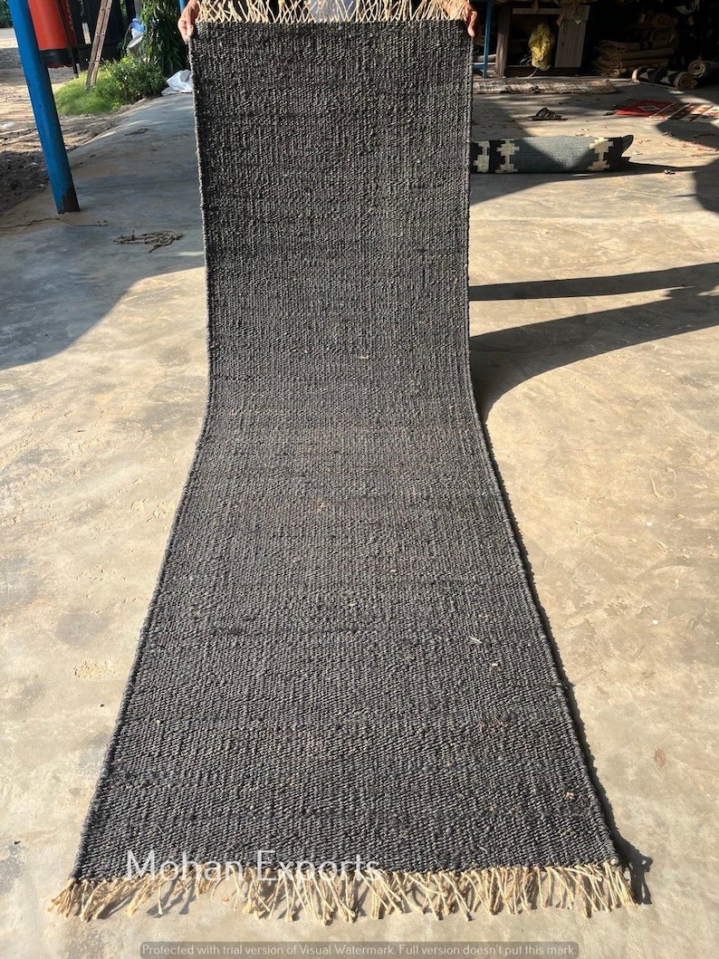 May include: A long, rectangular charcoal gray rug with a woven texture. The rug has a natural fiber fringe at the bottom edge. The rug is displayed outdoors on a concrete surface. The text "Mohan Exports" is visible.