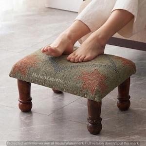 May include: A small, upholstered footstool with a patterned top in shades of green, orange, and blue. The footstool has four dark wooden legs. The text "Mohan Exports" is visible on the side of the footstool.
