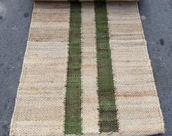 May include: A rolled-up jute runner rug with two vertical green stripes. The rug is made of natural fibers and has a woven texture. The text "Mohan Exports" is visible on the bottom left corner.