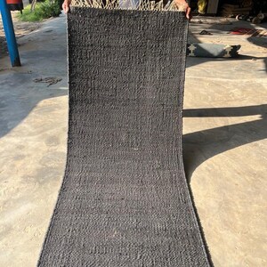 May include: A dark gray woven rug with a rectangular shape. The rug has a textured surface and a fringe detail at the top edge. The rug is held up by a person.