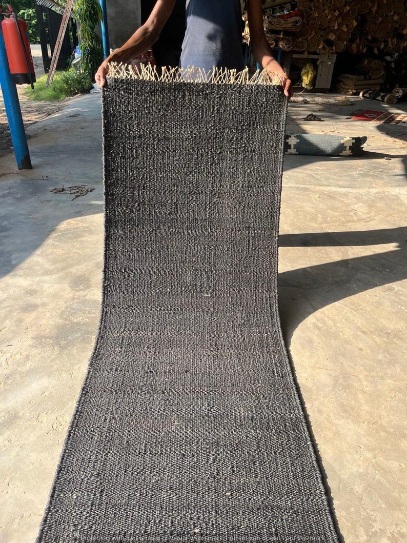 May include: A dark gray woven rug with a rectangular shape. The rug has a textured surface and a fringe detail at the top edge. The rug is held up by a person.
