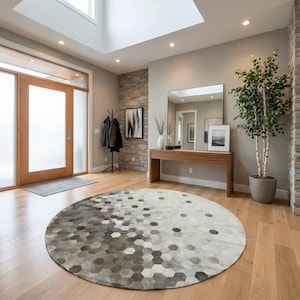 May include: A modern entryway with a large round rug featuring a geometric pattern of gray and white hexagons. The room has light wood flooring, a wooden console table with a mirror, and a potted tree. A wooden door and a stone accent wall are also visible.