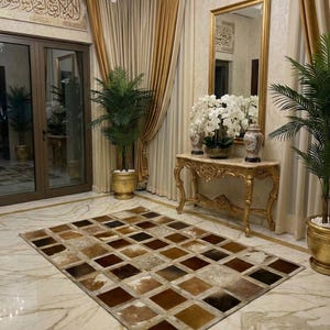 Op de afbeelding: Een rechthoekig vloerkleed met een patchworkontwerp van bruine, beige en crèmekleurige vierkanten. Het kleed ligt op een marmeren vloer in een kamer met gouden accenten, een spiegel en potplanten. De kamer heeft een luxueuze en elegante uitstraling.