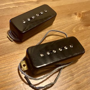 May include: Two black guitar pickups with silver screw heads and gray wiring are displayed on a wooden surface. The rectangular pickups have a glossy finish and are designed for electric guitars.