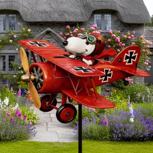 Puede incluir: Decoración de jardín de avión biplano rojo con un personaje de perro de dibujos animados que lleva un casco y gafas de piloto. El avión tiene cruces negras en las alas y la cola, una hélice de madera y ruedas negras. La decoración está en un jardín con flores y una casa de piedra.