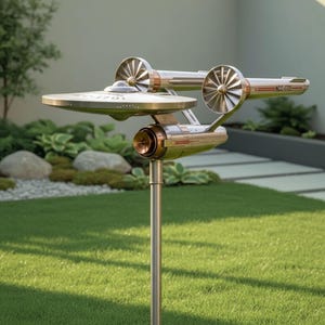 May include: A metal sculpture of the Starship Enterprise, mounted on a pole for garden display. The sculpture features a saucer section, warp nacelles, and the ship's registry number, NCC-1701. The sculpture is set against a backdrop of green grass and landscaping.