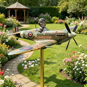 Può includere: Un modello dettagliato di un aereo da caccia della Seconda Guerra Mondiale, con una figura di pilota. L'aereo è dipinto con colori mimetici con testo Iron Maiden. Il modello è montato su un supporto dorato, in un giardino.
