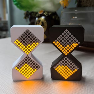 May include: Two hourglass-shaped digital clocks, one white and one black. Each clock features a grid of small squares that illuminate in yellow, creating a visual display. The clocks are designed with a modern aesthetic.
