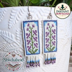 May include: A pair of rectangular beaded earrings featuring a floral pattern in purple, green, and white, framed by a blue border. The earrings hang from silver-toned hooks and include a fringe of white, blue, and purple beads at the base.