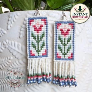 May include: A pair of rectangular beaded earrings featuring a floral motif in pink, green, and white, framed by a blue border. The earrings have long white fringe at the bottom, accented with small blue beads at the tips and silver-toned hooks.