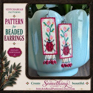 May include: A pair of rectangular beaded earrings featuring a ladybug and pink flower design on a cream background with a red border. The earrings have dangling bead fringe at the bottom and silver-toned metal hooks for attachment.