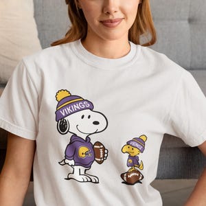 May include: A white short-sleeved t-shirt featuring a graphic of Snoopy and Woodstock wearing purple and yellow Vikings beanies and hoodies. Snoopy holds a brown football, while Woodstock stands on a second brown football. The text VIKINGS is printed on both beanies in white letters.