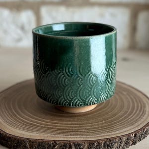 May include: A cylindrical ceramic cup with a glossy dark green glaze. The lower portion of the exterior features a repeating embossed pattern of overlapping semicircular waves. The base of the cup is unglazed, revealing a light tan clay color.