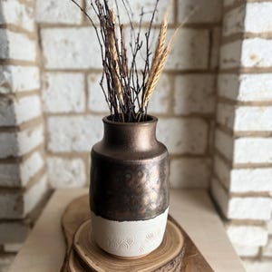 May include: A ceramic vase with a metallic bronze upper section and a textured white lower section featuring a repeating wave pattern. The vase contains a collection of thin dark brown twigs and golden wheat stalks arranged vertically inside.