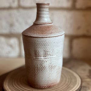 May include: A cylindrical ceramic bottle with a narrow neck, featuring a textured off-white glaze with visible reddish-brown clay undertones. The body is decorated with a repeating embossed leaf-like pattern and horizontal bands around the neck.