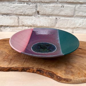 May include: A handmade ceramic bowl with a unique color scheme. The bowl features sections of pink, purple, and teal, with a dark blue center. The bowl is placed on a wooden surface, showcasing its handcrafted nature and artistic design.