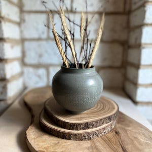 May include: A round, sage green ceramic vase with a smooth, matte finish and a narrow neck. The vase contains several thin, dried brown twigs and stalks of wheat. It sits atop two stacked circular wood slices with visible bark edges.