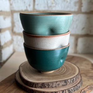 May include: A stack of three ceramic bowls with a dimpled texture. The top bowl is light sage green, the middle bowl is off-white with speckles, and the bottom bowl is a deep forest green. Each bowl features a raw, unglazed brown clay rim.