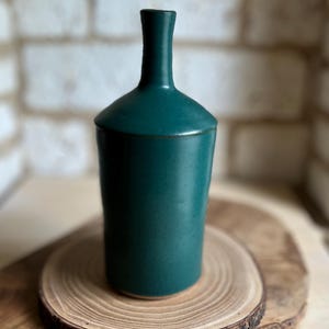 May include: A dark teal ceramic bottle with a smooth, matte finish, featuring a cylindrical body that tapers into a narrow, slender neck. The base of the bottle is slightly wider than the top, showcasing a simple, clean, and functional design.