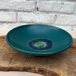 May include: Teal-colored ceramic bowl with a speckled finish. The center features a circular design with shades of blue and green. The bowl is set on a wooden surface, showcasing its unique shape and color.