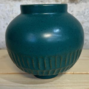 May include: Teal-colored ceramic vase with a rounded body and a narrow neck. The vase features a textured, ridged design around the base. The surface has a speckled appearance. The vase is displayed on a light-colored wooden surface.