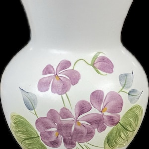 Puede incluir: Jarrón de cerámica blanca con cuerpo bulboso y parte superior acampanada. El jarrón está decorado con flores moradas pintadas a mano, hojas verdes y centros amarillos. El jarrón mide aproximadamente 20 cm de altura.