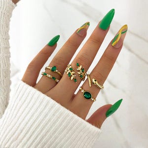 Puede incluir: Una mano que muestra una colección de anillos dorados con piedras verdes y diseños de hojas. Los anillos incluyen piedras con corte esmeralda y ovalado. Las uñas están pintadas de verde brillante con detalles dorados, complementando las joyas.