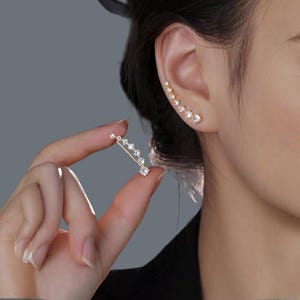 Puede incluir: Un pendiente de oreja dorado y un trepador de oreja a juego, ambos adornados con brillantes cristales transparentes. El pendiente de oreja se sostiene en una mano, mostrando el delicado diseño. El trepador de oreja se lleva en la oreja.