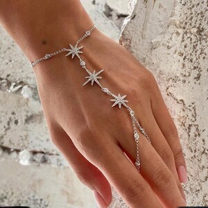 May include: A delicate silver hand chain featuring a series of star-shaped charms adorned with sparkling crystals. The bracelet wraps around the wrist and connects to a ring, creating a celestial design. The jewelry is a beautiful accessory.